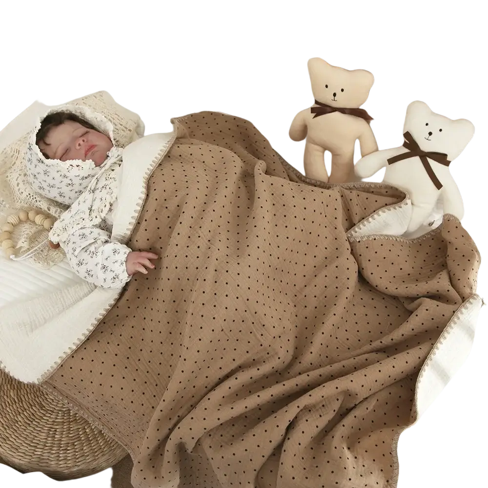 Muslin cotton baby swaddle blanket in beige with black polka dots and white border draped over a sleeping baby in a crib