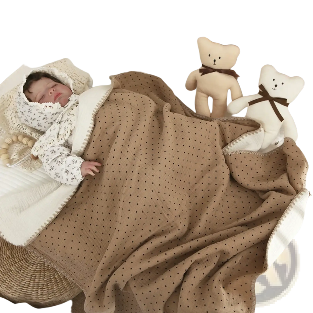 Muslin cotton baby swaddle blanket in beige with black polka dots and white border draped over a sleeping baby in a crib