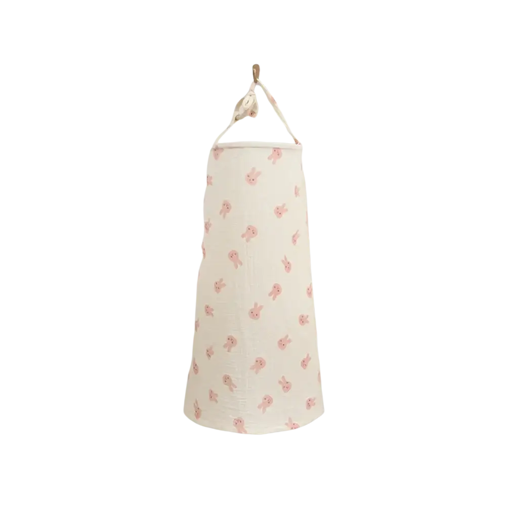 A cream cotton laundry hamper with pink bunny pattern hanging on wall paired with a breathable nursing cover dinosaur print