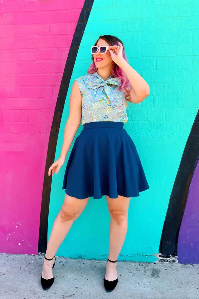 A woman wearing a sleeveless floral blouse with bow paired with an ocean blue high waisted pleated skirt with pockets