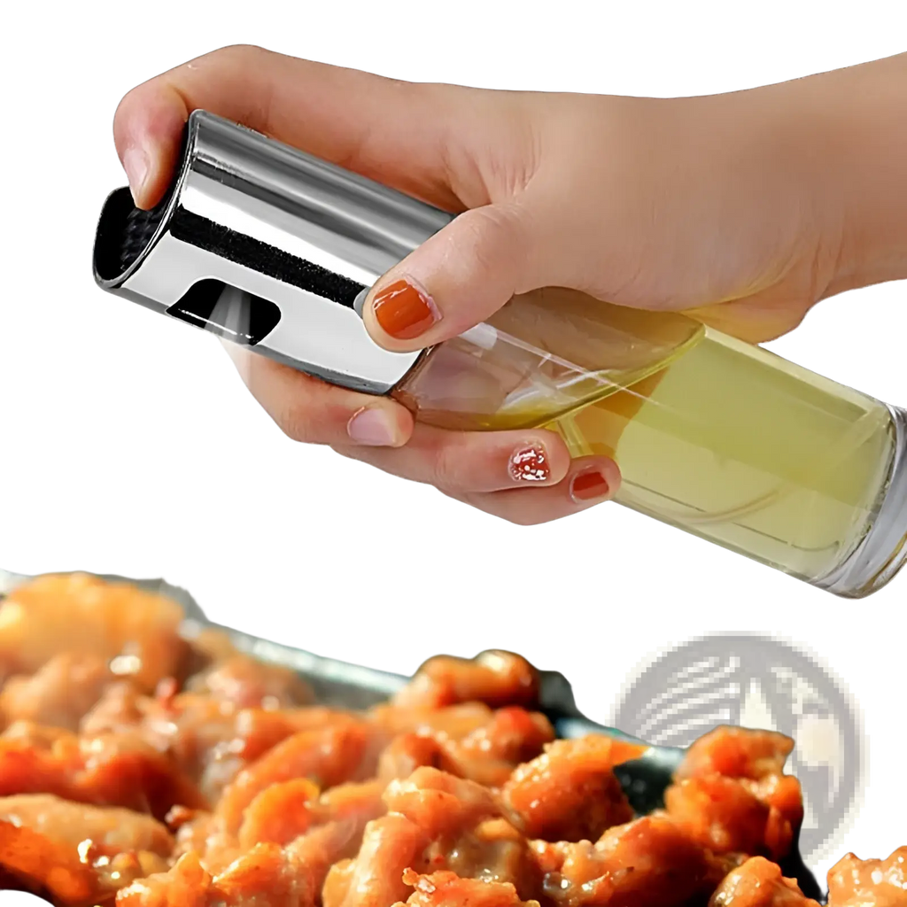 A sleek transparent glass oil spray bottle with a chrome nozzle and black trigger misting oil over cooking food