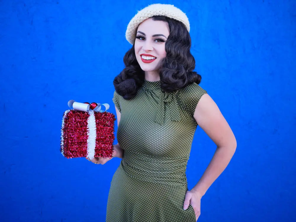 A festive red and white tinsel-wrapped gift box with a silver ribbon bow alongside an olive dot bombshell retro plus size flare dress