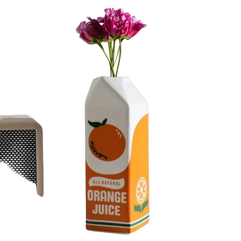 A white and orange ceramic vase shaped like a carton with an all natural orange juice label and orange illustration