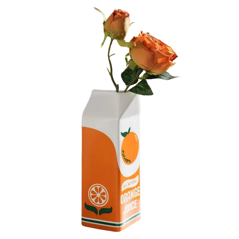 A white and orange ceramic vase shaped like an orange juice carton with an all natural orange juice label and orange slice illustration