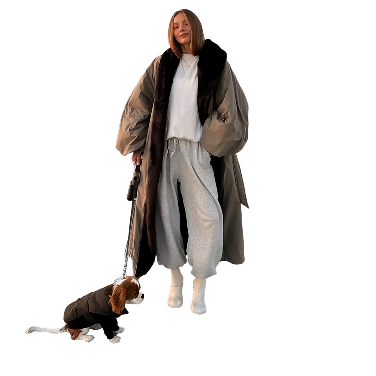 A woman wearing an oversized beige trench coat with a dark brown fur-lined hood stands on a paved promenade