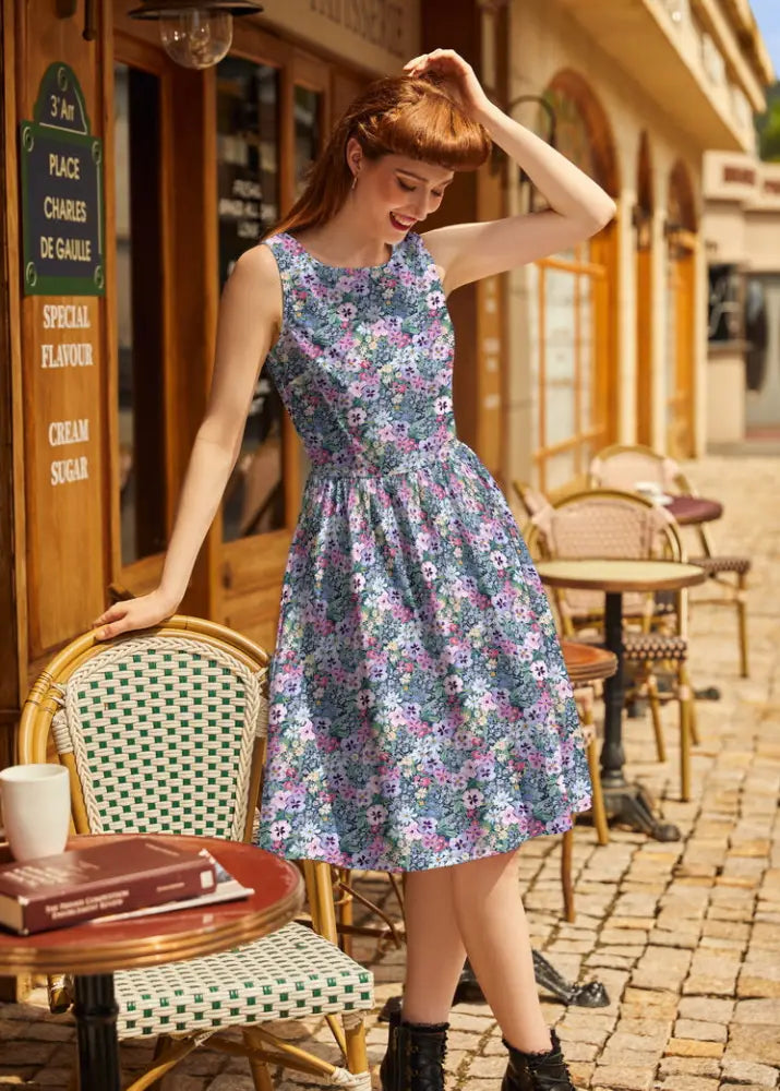 A woman wears a sleeveless 1950s painted floral cotton a-line midi dress with blue and purple flowers and pockets