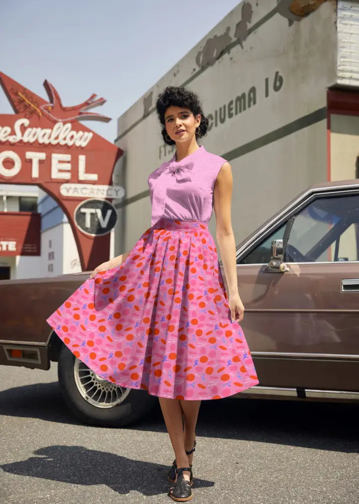 A woman wearing a sleeveless lavender top with a bow pairs it with a pink cotton Paris Doris retro pleated skirt with pockets