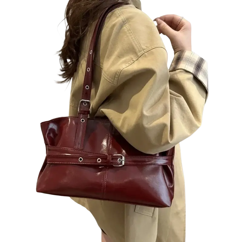 A glossy burgundy patent leather shoulder bag with adjustable strap silver buckle straps and decorative metal eyelets
