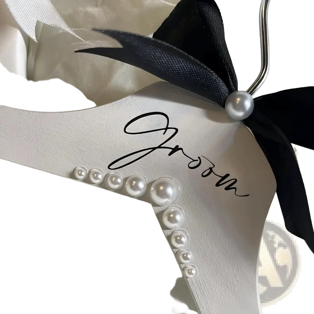White wooden groom hanger with black ribbon bow, pearl accent, and cursive lettering adorned with graduated pearls