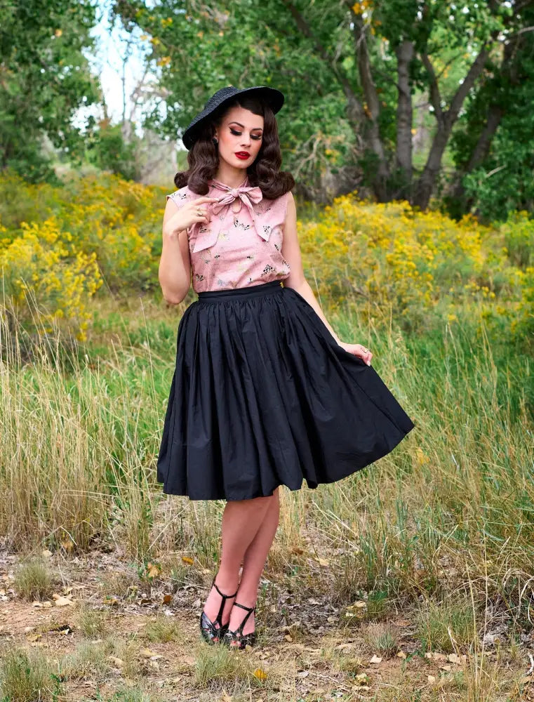 A woman wearing a pink floral blouse with bow paired with a black Peggy black 1950s cotton swing skirt with pockets