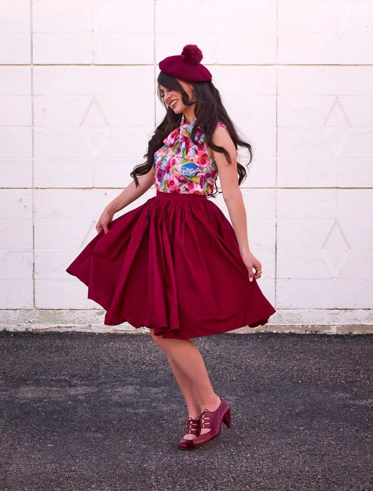 A woman wearing a vibrant floral top with a rich burgundy Peggy cotton 1950s swing skirt with pockets and beret