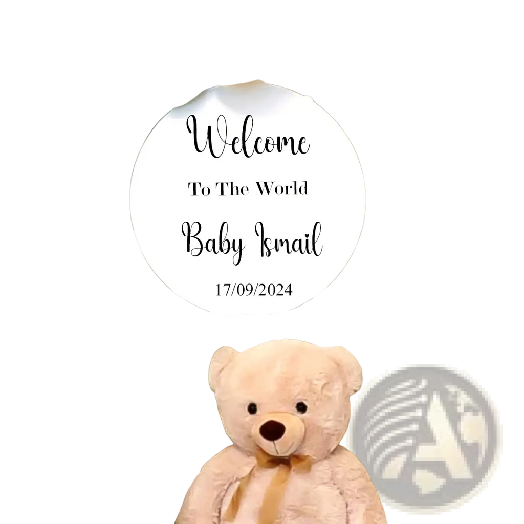 A plush light beige teddy bear with tan ribbon sits in front of a personalized round baby shower welcome sign kt board