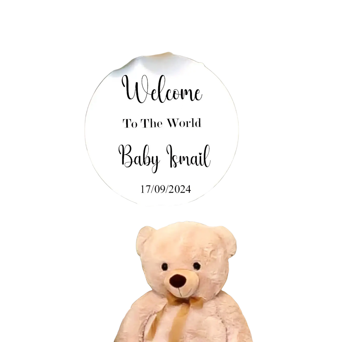 A plush light beige teddy bear with tan ribbon sits in front of a personalized round baby shower welcome sign kt board