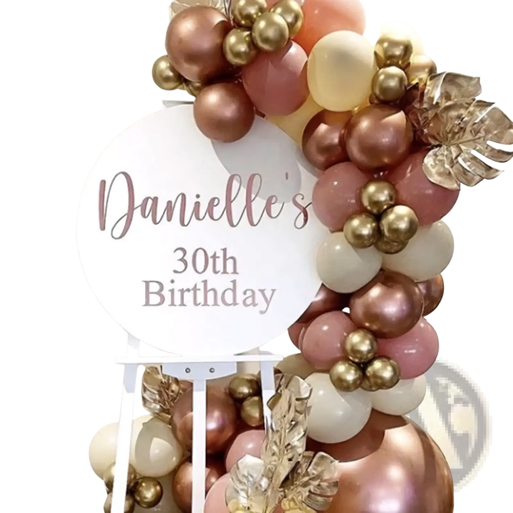 Personalized round baby shower welcome kt board with a rose gold and cream balloon arch accented by metallic leaves