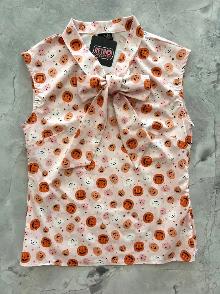 Sleeveless light pink blouse with orange and pink smiling pumpkins and stars, featuring a large bow at the collar