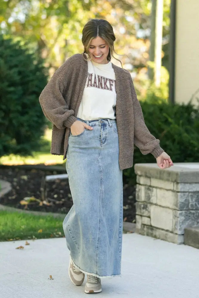 A woman wearing a piper light wash a-line denim maxi skirt with frayed hem paired with a brown knit cardigan