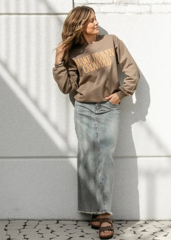 A woman wears a soft brown long-sleeved sweatshirt with gold lettering paired with a piper light wash denim maxi skirt