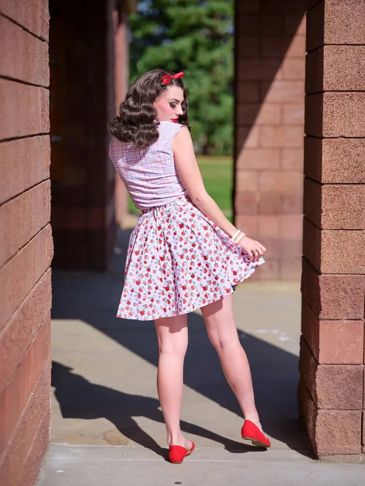 A woman wears a vintage pink plaid sleeveless bow blouse with bust darts and a flared skirt with red strawberry prints