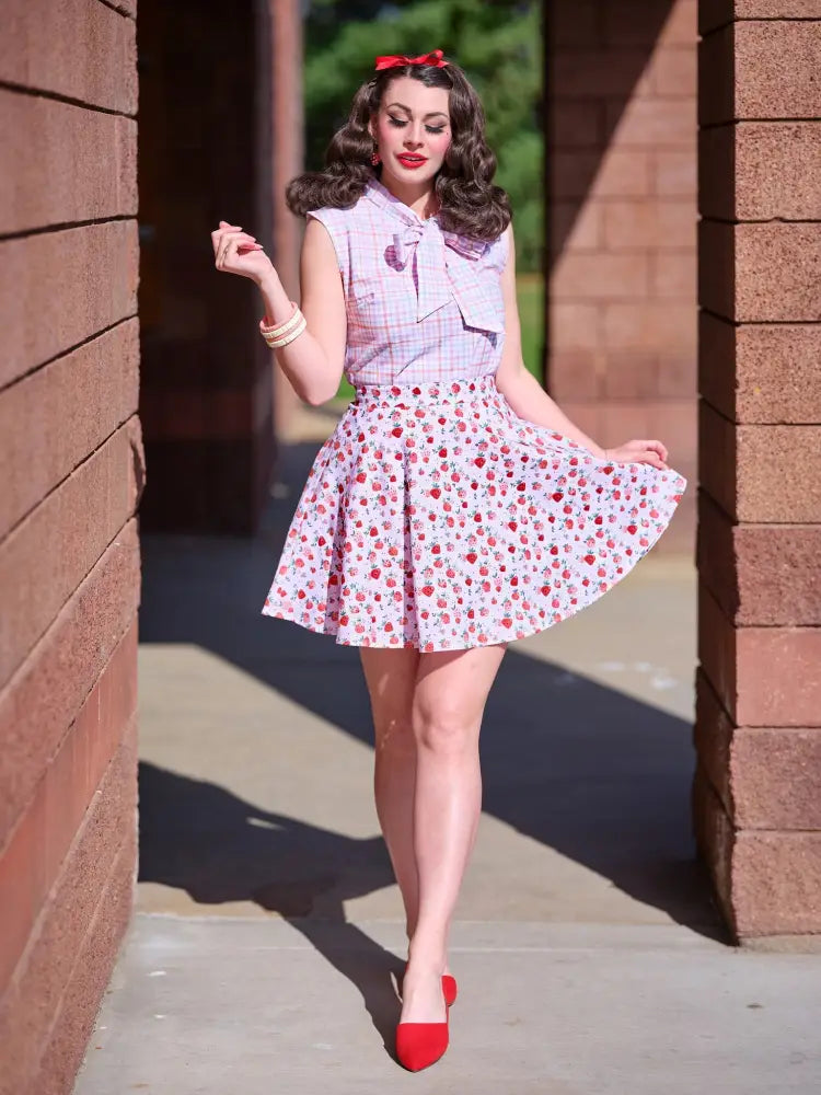 A woman wearing a vintage pink plaid sleeveless bow blouse with bust darts paired with a cherry print skirt