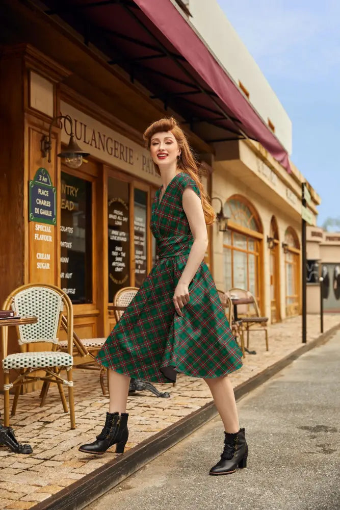 A woman wearing a green plaid v neck cotton a line dress with short sleeves, fitted waist, and pockets