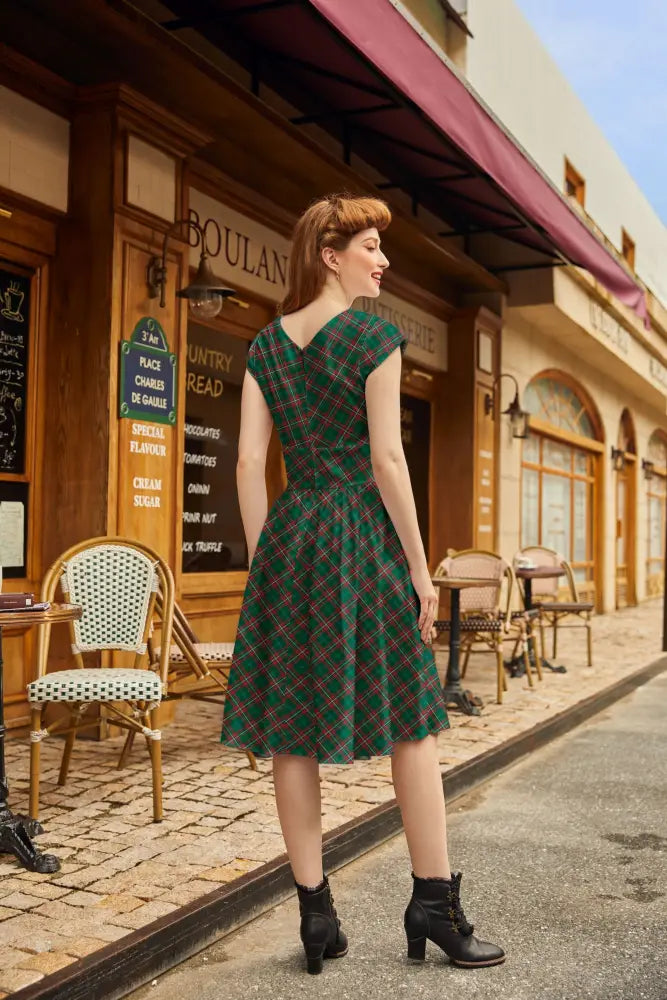 A woman wearing a green and red plaid cotton a line dress with v neck, fitted waist, short sleeves, and pockets