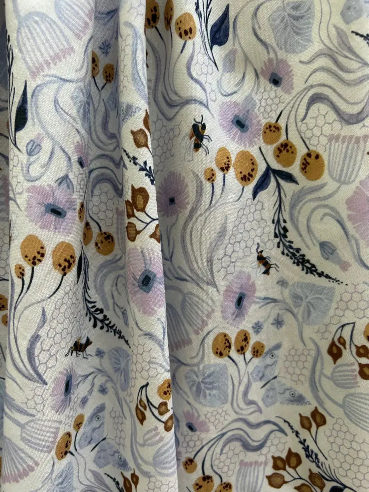 A cream cotton a-line dress with pockets featuring a vintage pollinators floral and insect pattern in muted purples golds and blues