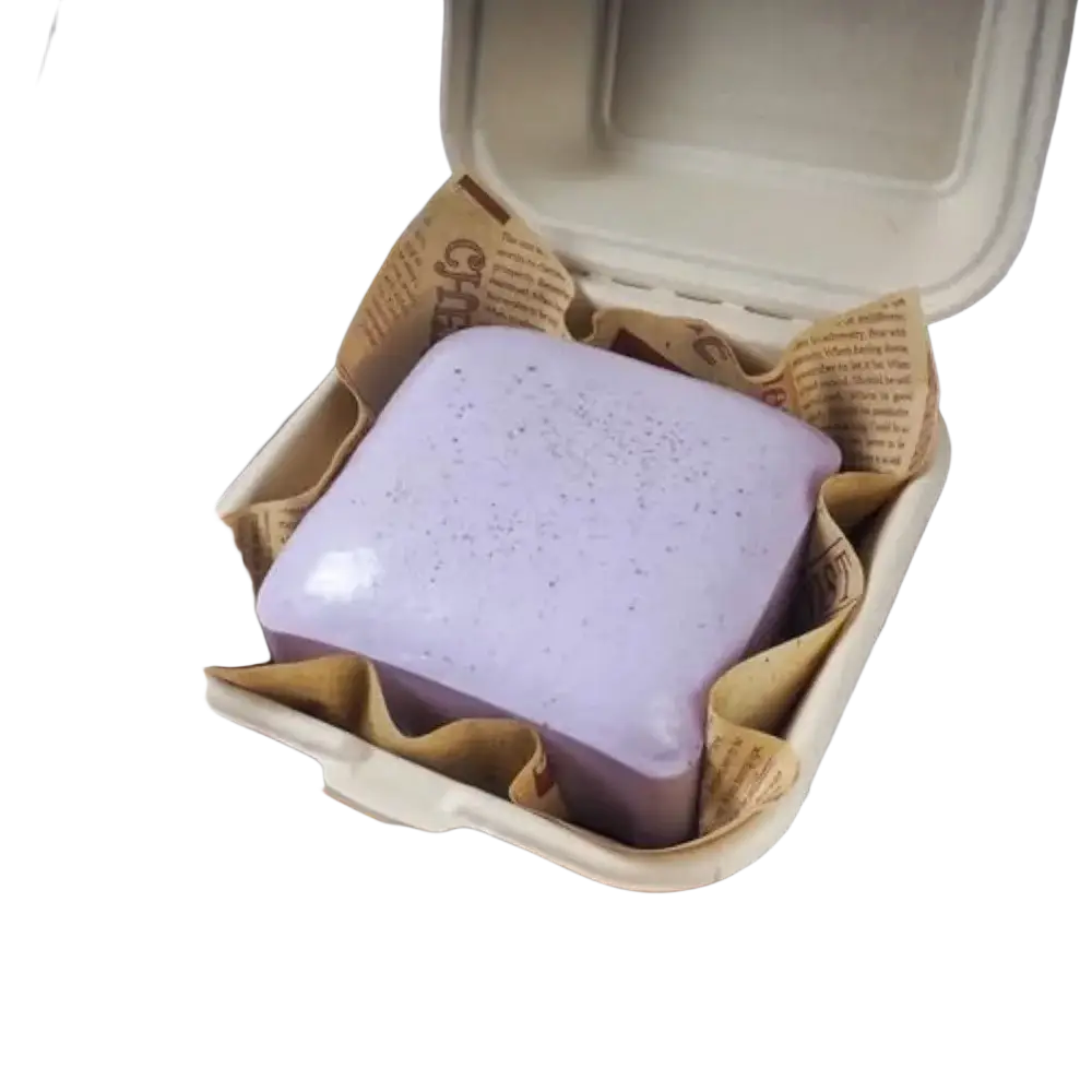 A lavender square-shaped soap bar with speckled texture in a paper-lined container alongside a polyester acrylic winter scarf