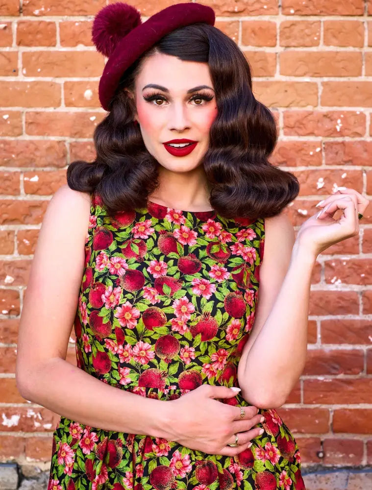A woman wears a pomegranate floral 1950s cotton swing midi dress in red and green with a maroon beret and pom-pom
