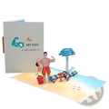A handmade pop up fathers day card featuring a muscular man in red swim trunks on a beach with umbrella and chairs