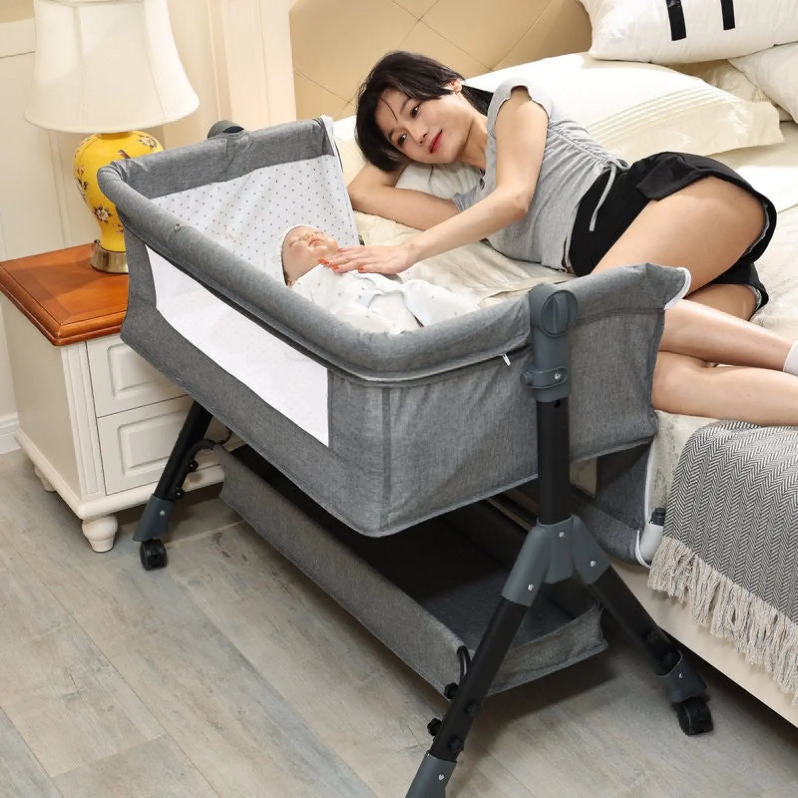 Gray fabric portable folding baby bassinet with black metal frame wheels mosquito net and mattress shown from the back