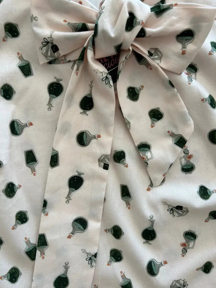 White fabric vintage sleeveless bow top with dark green potion bottle print and side zipper detail tied into a bow