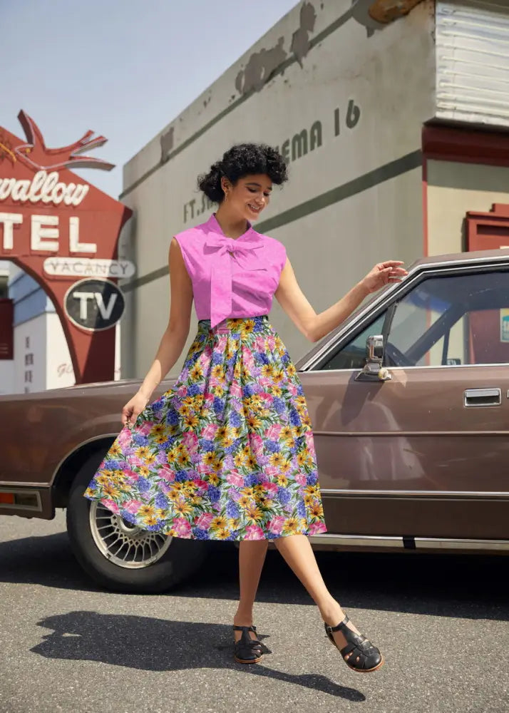 A woman wearing a retro pink mosaic cotton sleeveless top with bow neckline paired with a floral patterned skirt