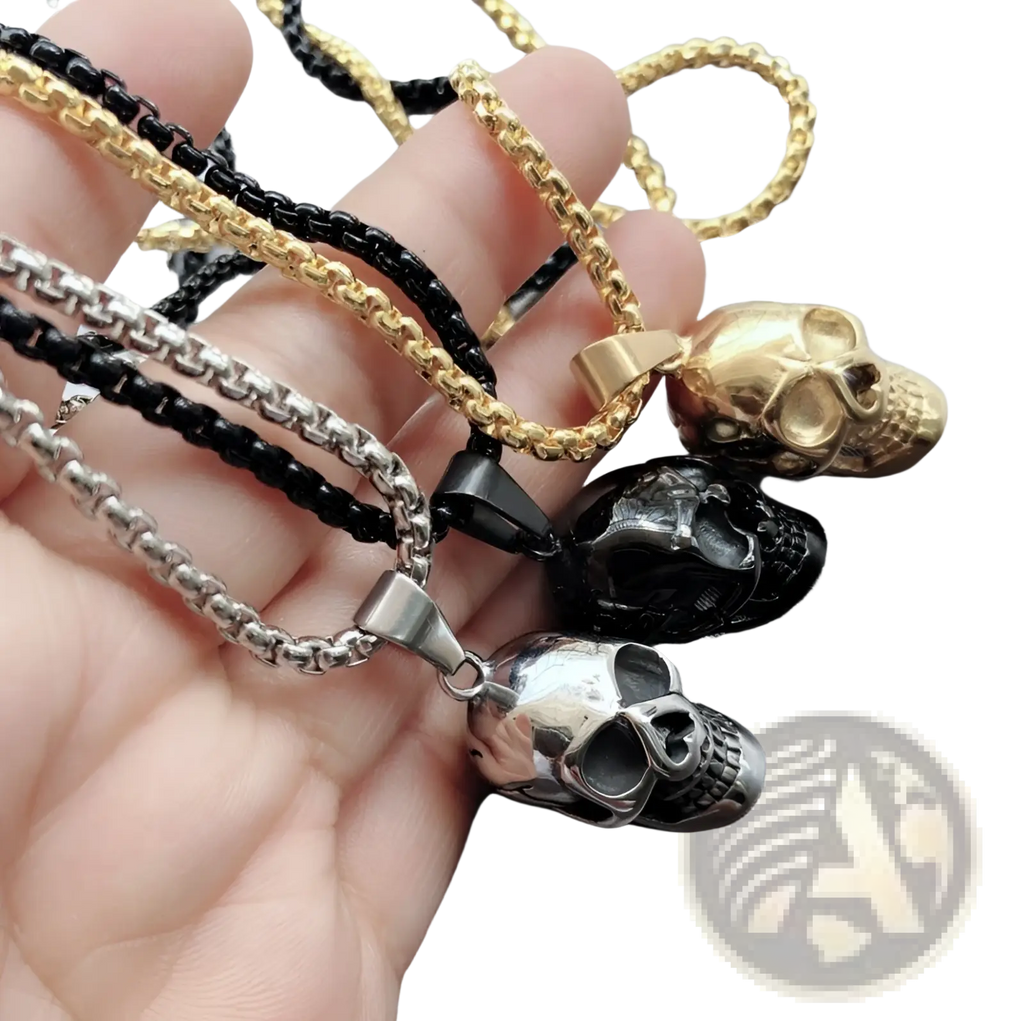 A hand holds three punk skull pendant titanium steel necklaces in gold, blackened steel, and polished silver finishes
