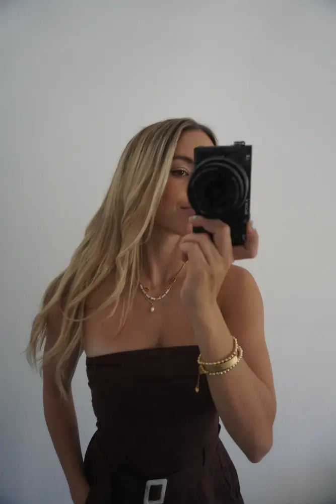 Black compact camera with prominent lens and minimalist design shown alongside pure pearl and gold bead layered necklace set