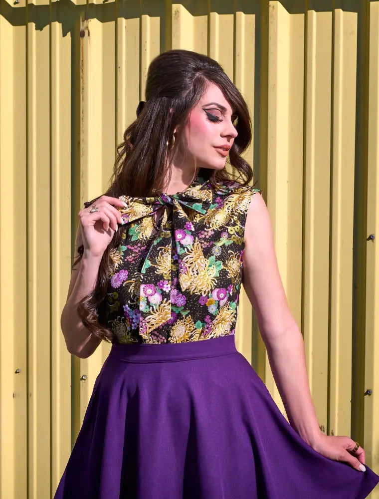 A vibrant floral-patterned sleeveless top with a black base and a bow paired with a purple pleated high waisted skater skirt with pockets