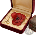 A glossy red enamel flower brooch with layered petals and black gemstone center in an elegant velvet box