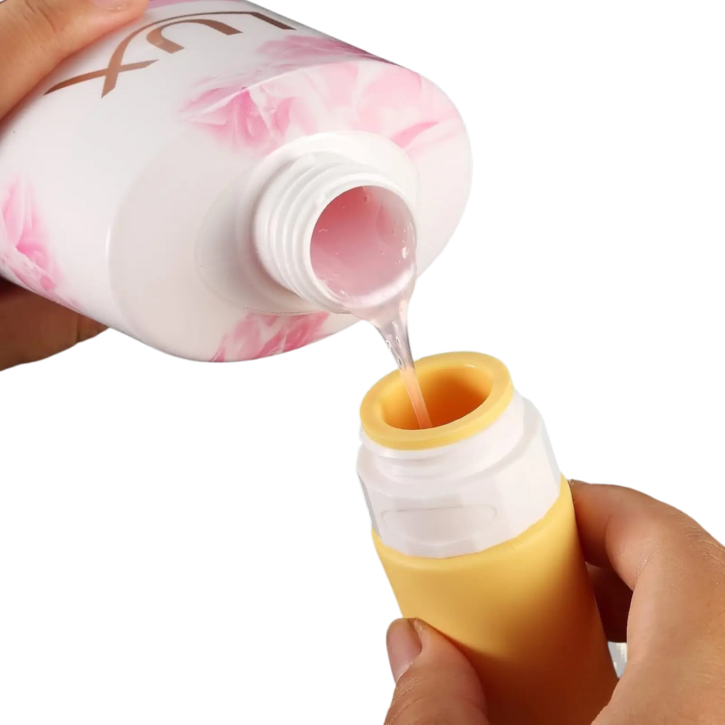 A white bottle with pink floral design and gold lettering pouring into a yellow refillable silicone travel bottle leakproof set
