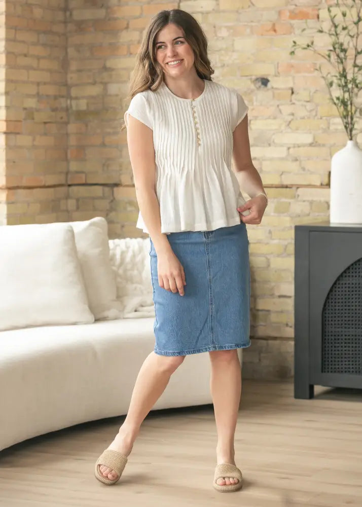 A woman wears a cream blouse with pleats and buttons paired with a medium-wash Remi classic wash stretch denim midi skirt
