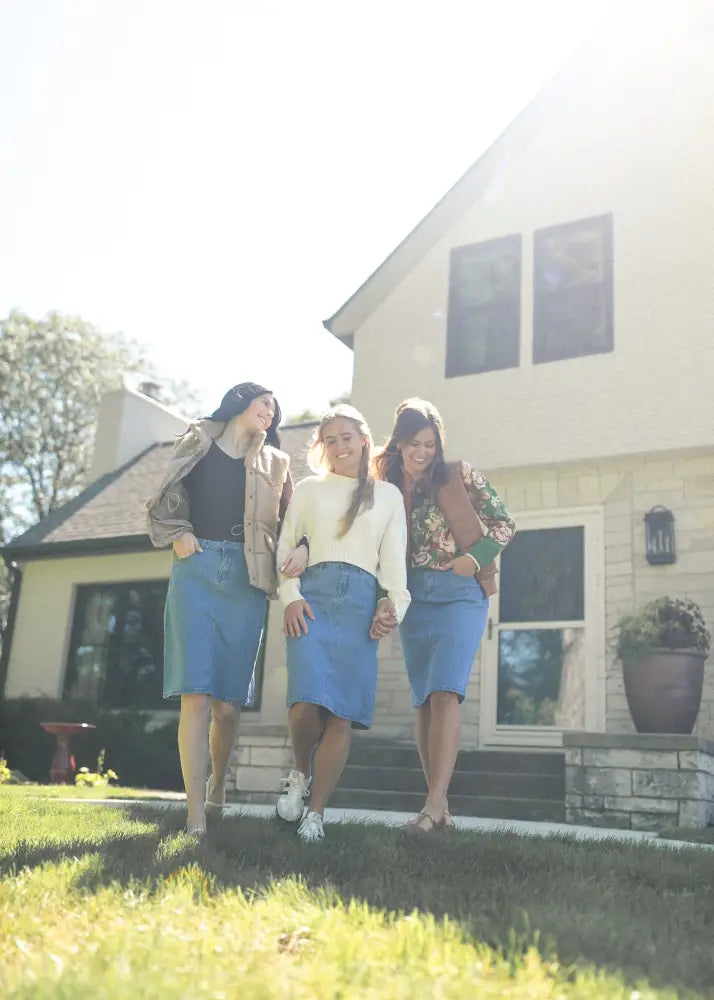 Three women walk together outside a house wearing remi classic wash stretch denim midi skirts in blue