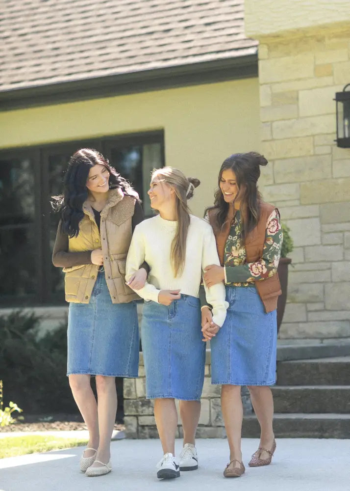 Three women wearing remi classic wash stretch denim midi skirts in blue denim walk together outside a house