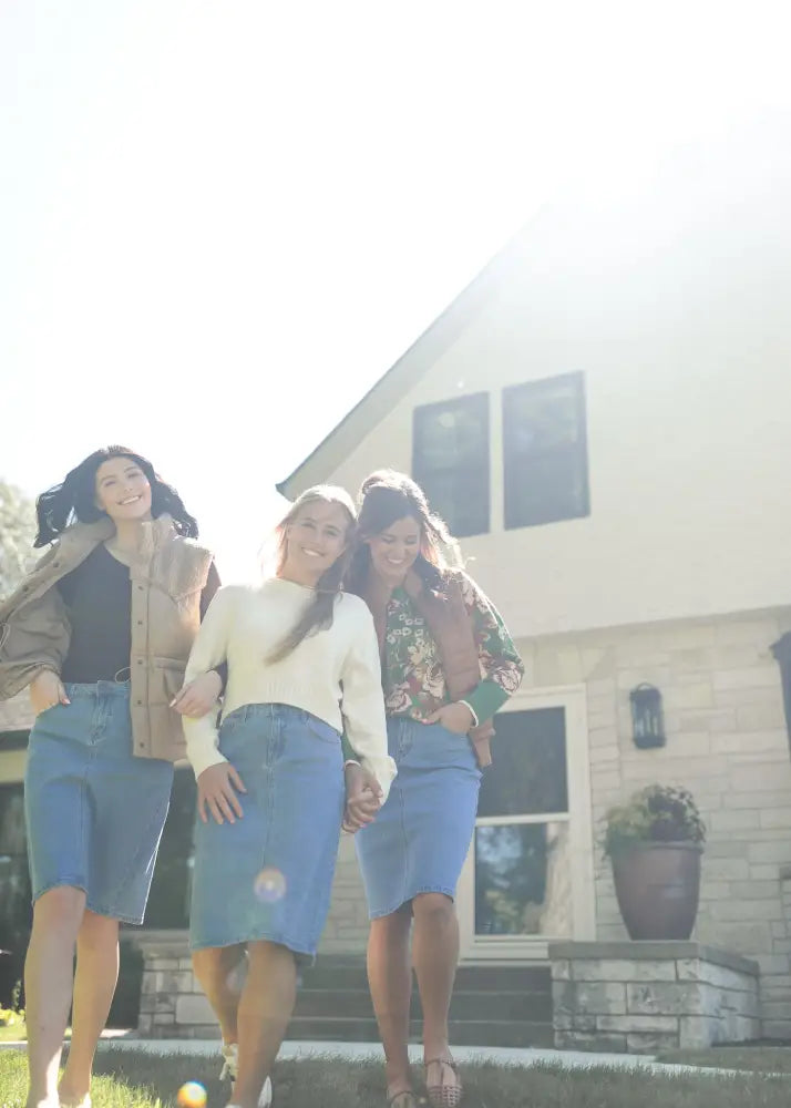 Three women walk outside a house wearing matching Remi classic wash stretch denim midi skirts in blue