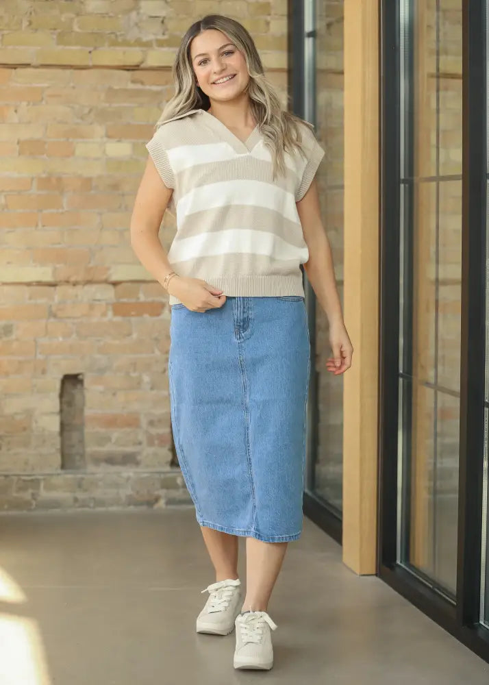 A woman wears a beige and white striped sleeveless knit vest over a blue remi classic wash stretch denim midi skirt