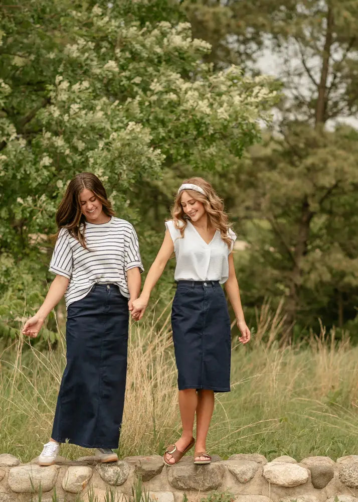Two women walk hand-in-hand one wearing a Remi navy high waisted stretch denim midi pencil skirt in front view
