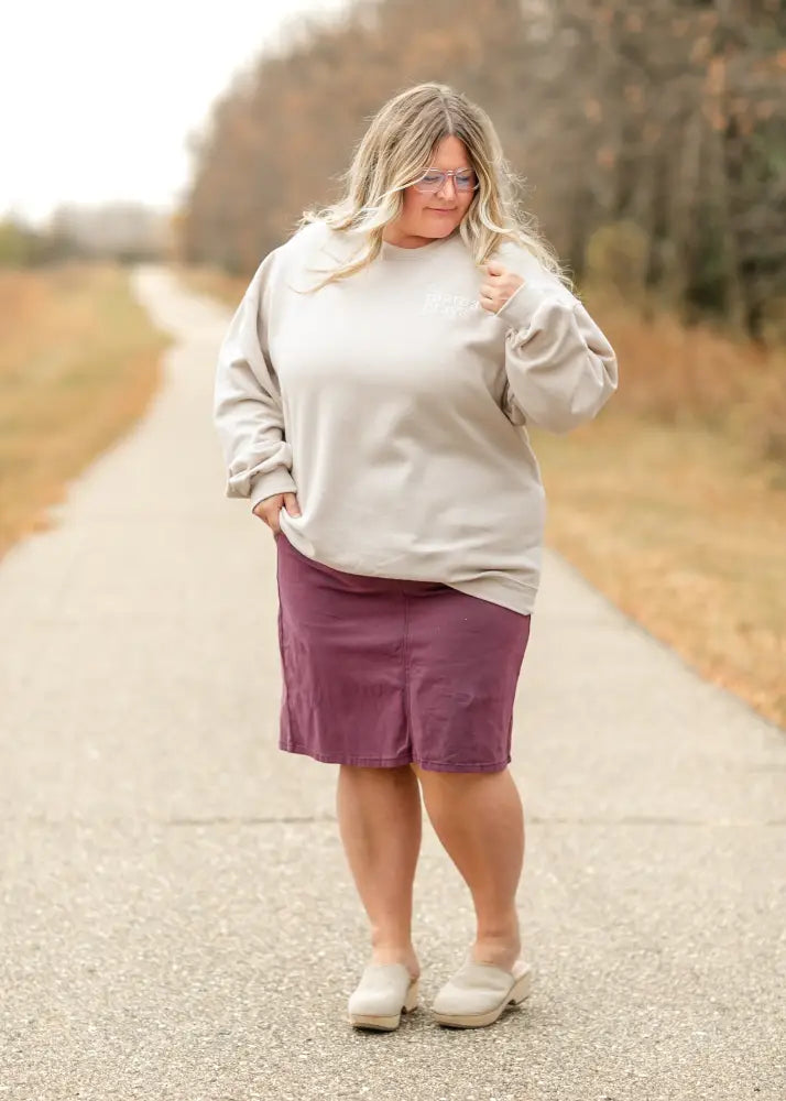A plus-size woman wearing a solid purple Remi Orchid burgundy denim midi skirt paired with a light gray sweatshirt