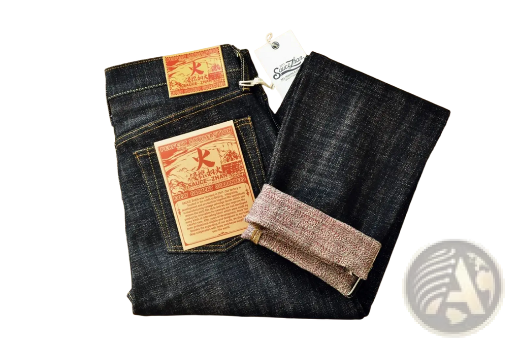 Dark indigo Saucezhan Fire Selvedge tapered raw denim jeans with red and orange branded patches folded on wood surface