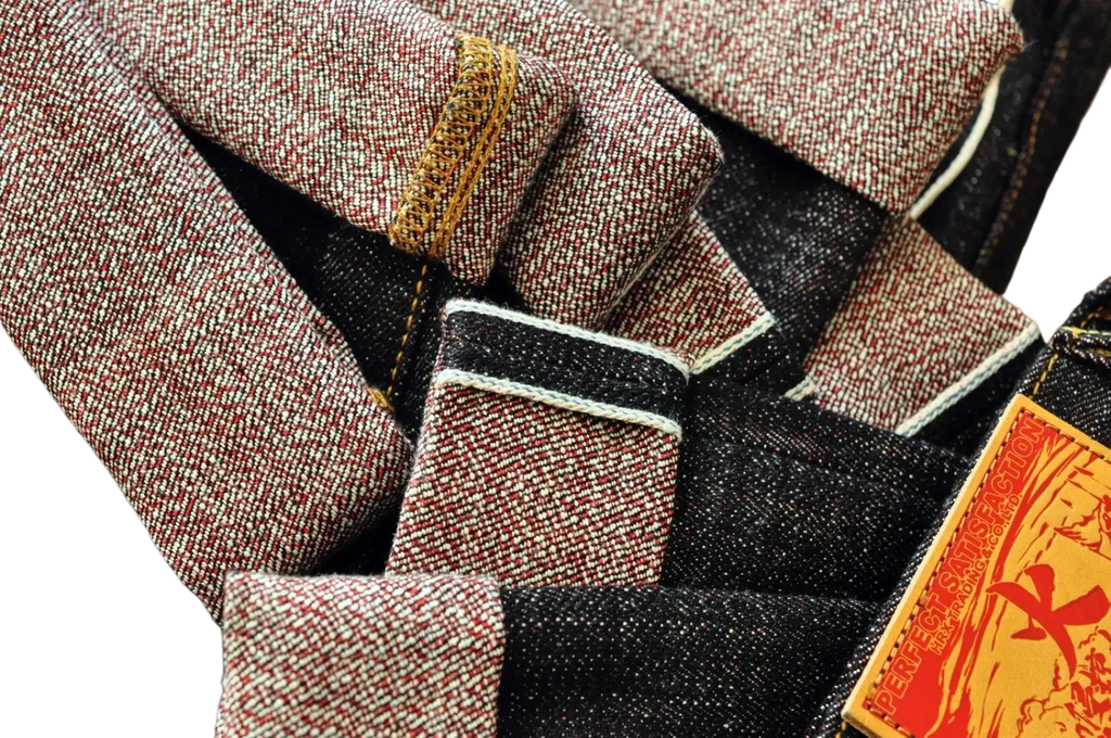A pair of dark denim Saucezhan fire selvedge tapered raw jeans with red and white speckled cuffs and yellow perfect selection label