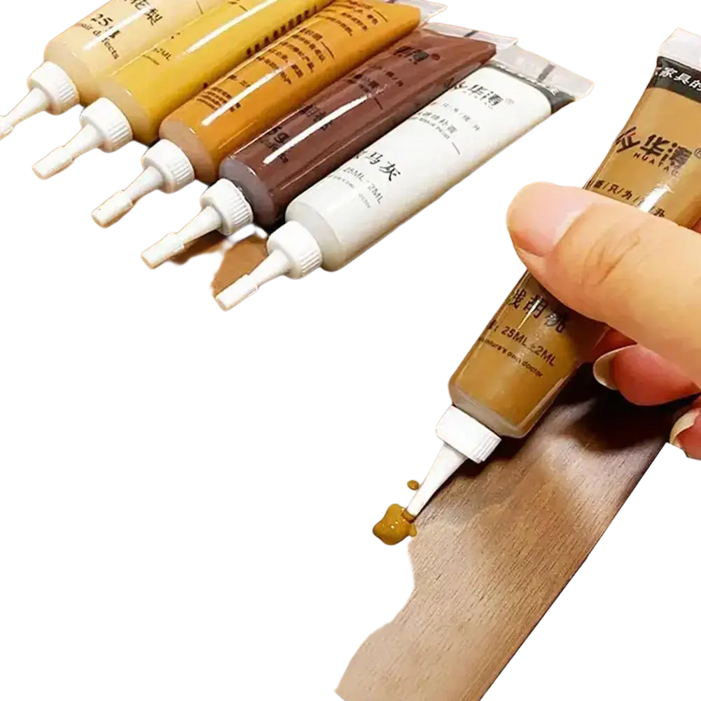 A hand holds a tube of brown repair paste from a touch-up paint kit with complementary paint tubes on wood