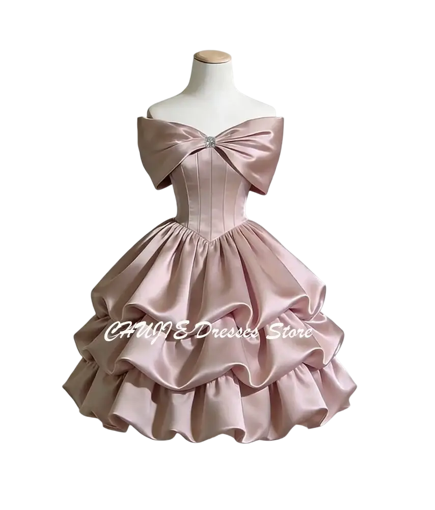 A blush pink satin off-shoulder graduation dress with a structured bodice, tiered ruffle skirt, and large bow neckline