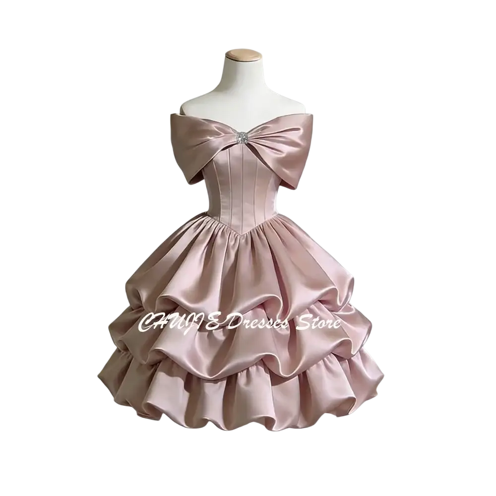 A blush pink satin off-shoulder tiered graduation dress with a structured bodice and large bow at the neckline