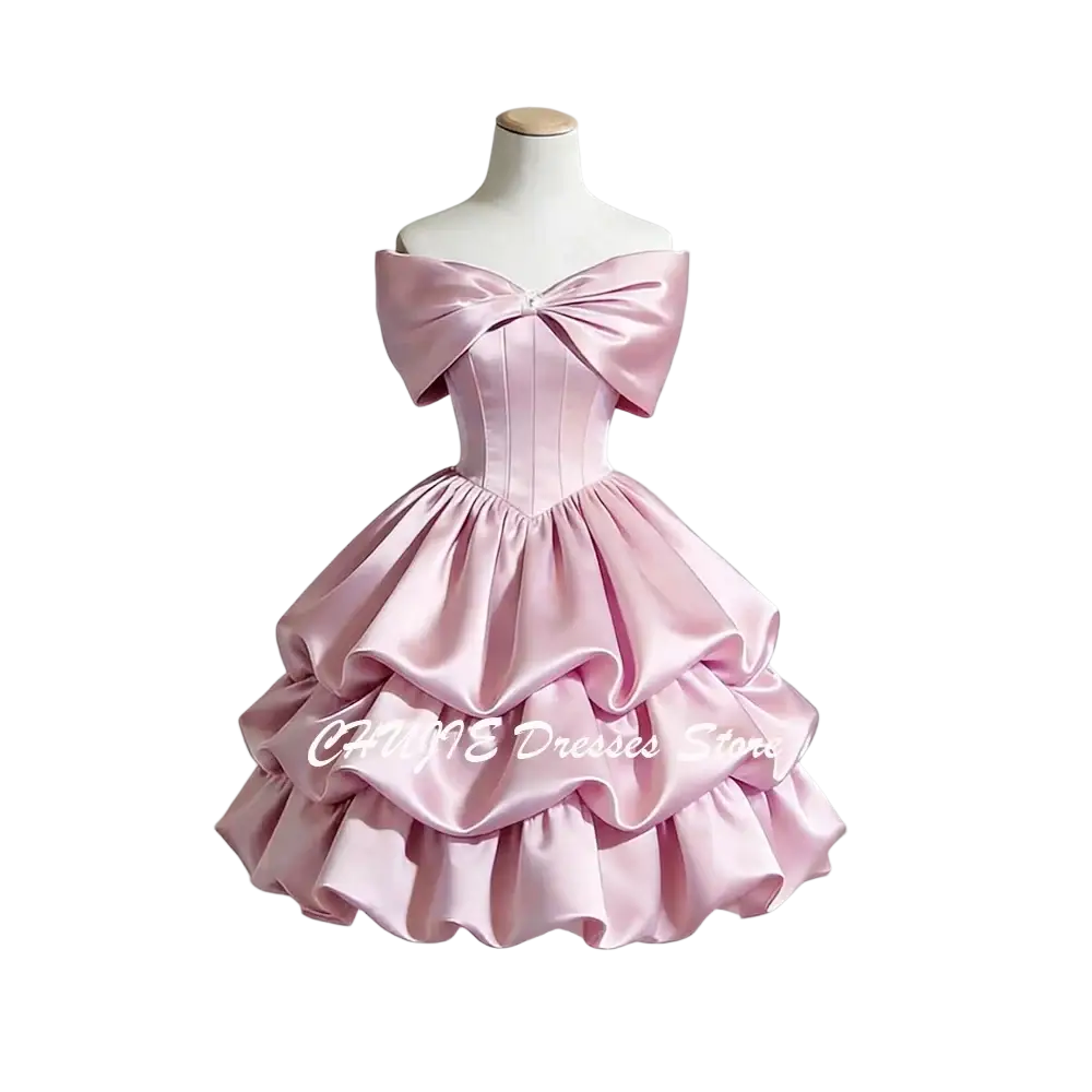 A delicate pink satin off-shoulder graduation dress with tiered ruffles and a large bow at the neckline